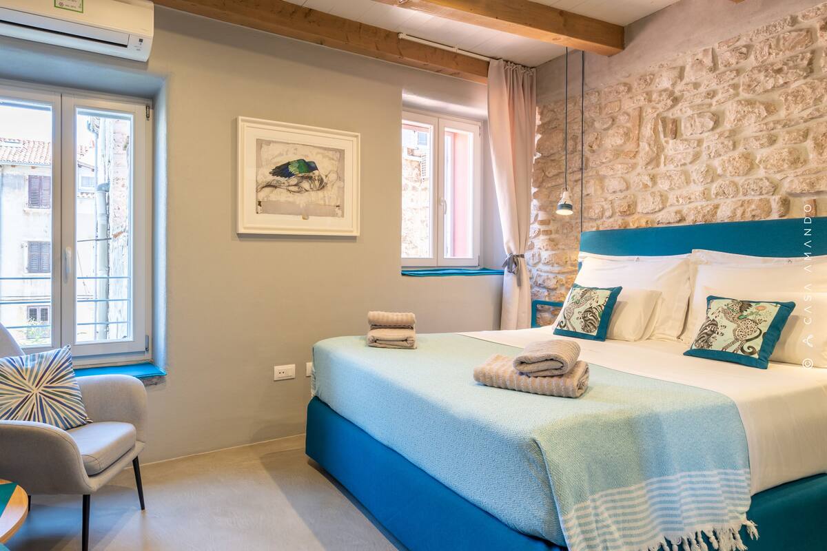 A bedroom with a bed, a chair, a table with a plant and fruit, and a stone wall.