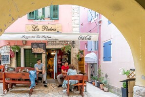 An outdoor café with wooden benches and tables, a pizzeria sign, and a Coca-Cola sign.