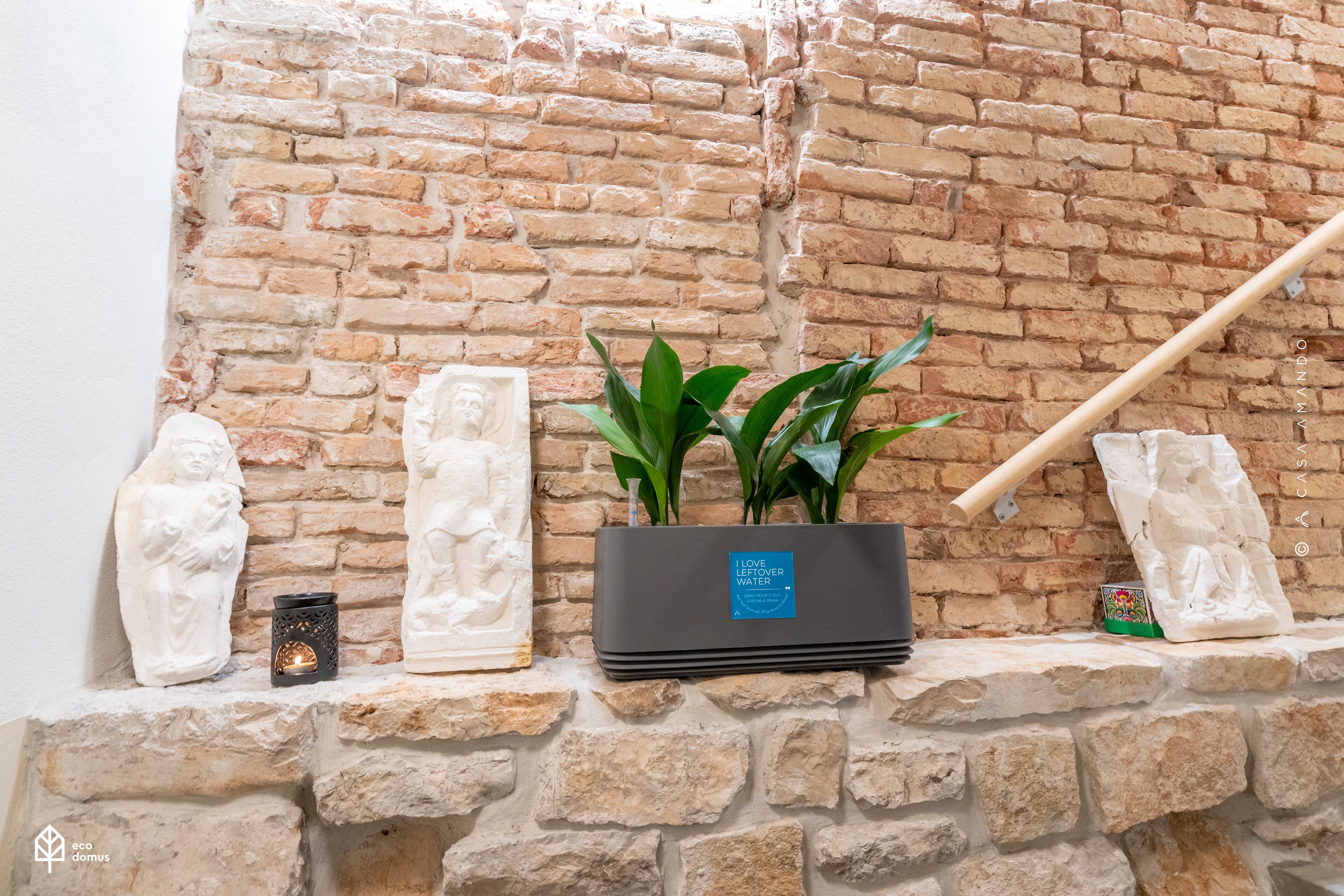 A stone wall with a brick pattern, a black planter with green plants, and white sculptures.