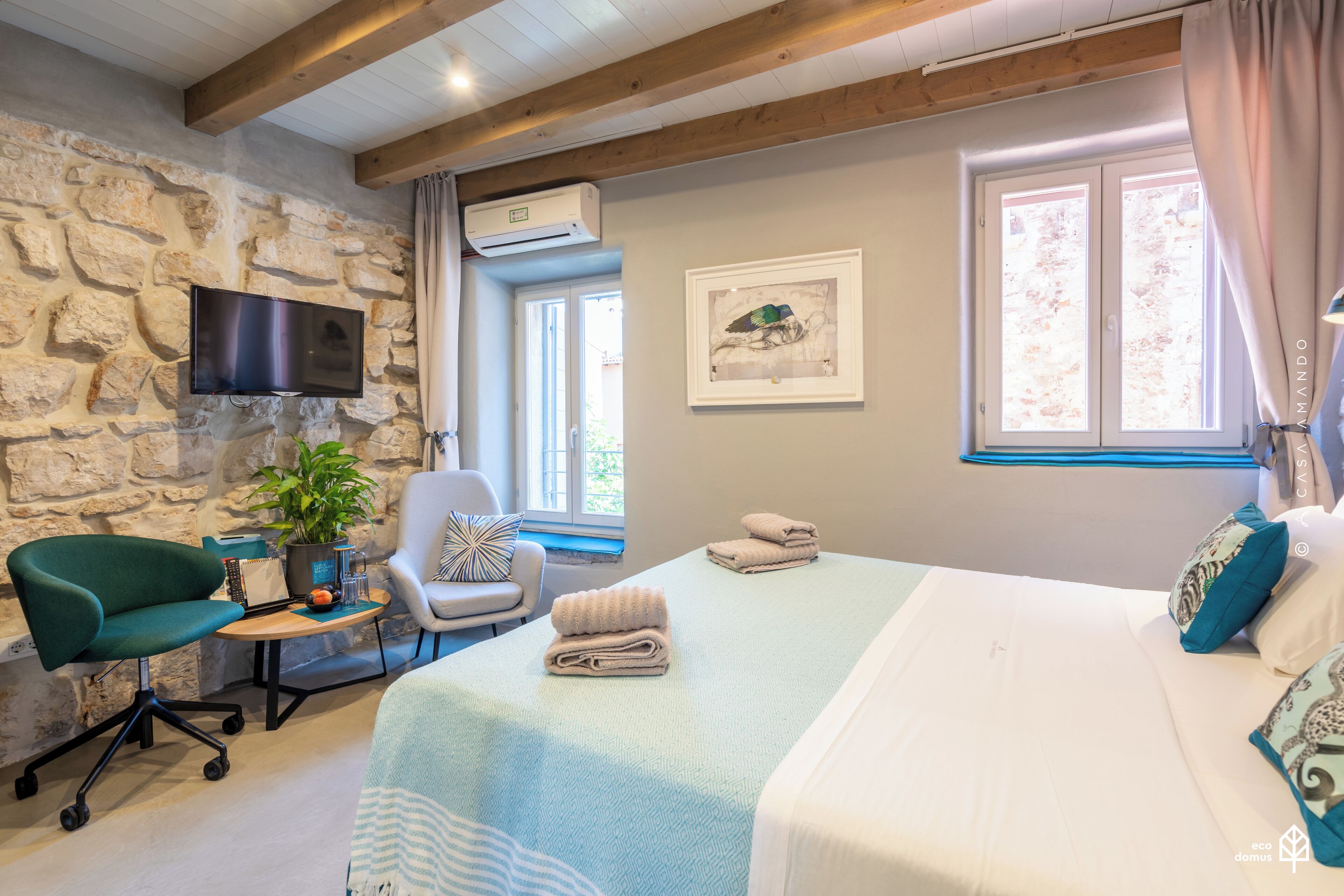 A bedroom with a stone wall, a bed with white bedding, a desk with a chair, a television, and a window with curtains.