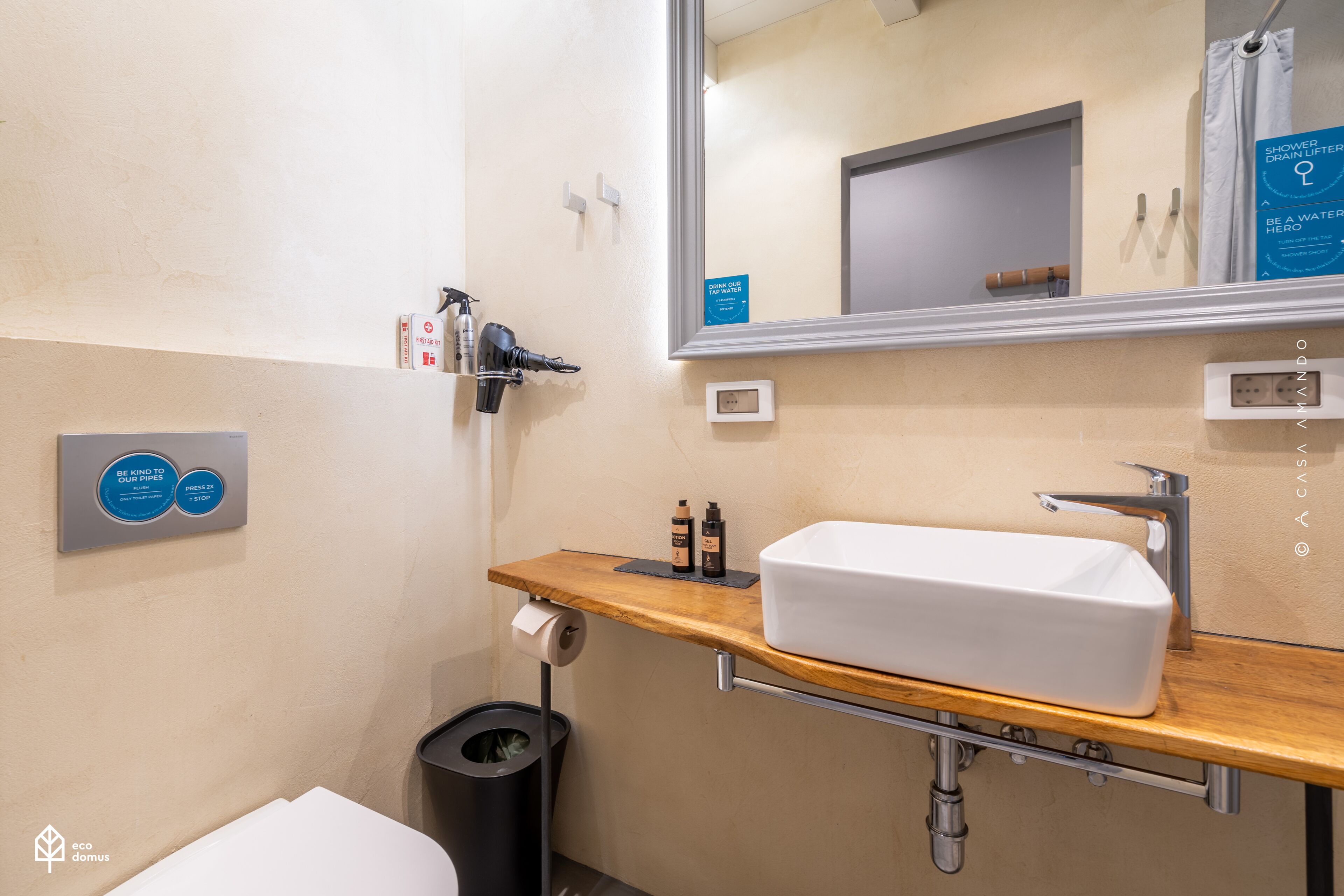 Une salle de bain moderne avec un meuble-lavabo en bois, un lavabo blanc et un miroir. On y trouve des articles de toilette sur le meuble-lavabo, une poubelle et un panneau encourageant à économiser l’eau.