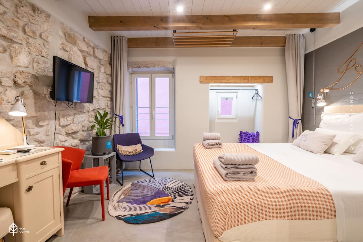 A modern hotel room with a stone wall, a flat-screen TV, a desk with a lamp, a red chair, a bed with an orange bedspread, and a window with curtains.