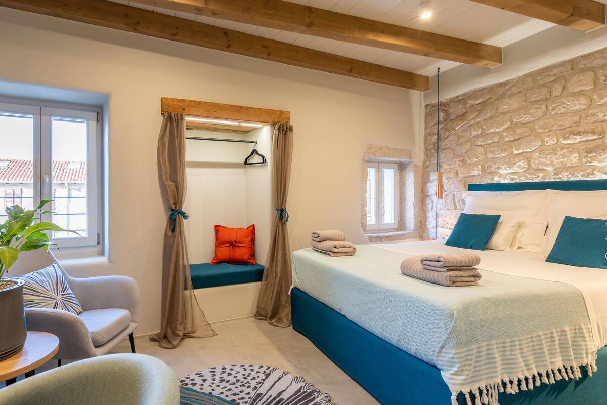A bedroom with a bed, a seating area with a chair and a table, a potted plant, and a stone wall.