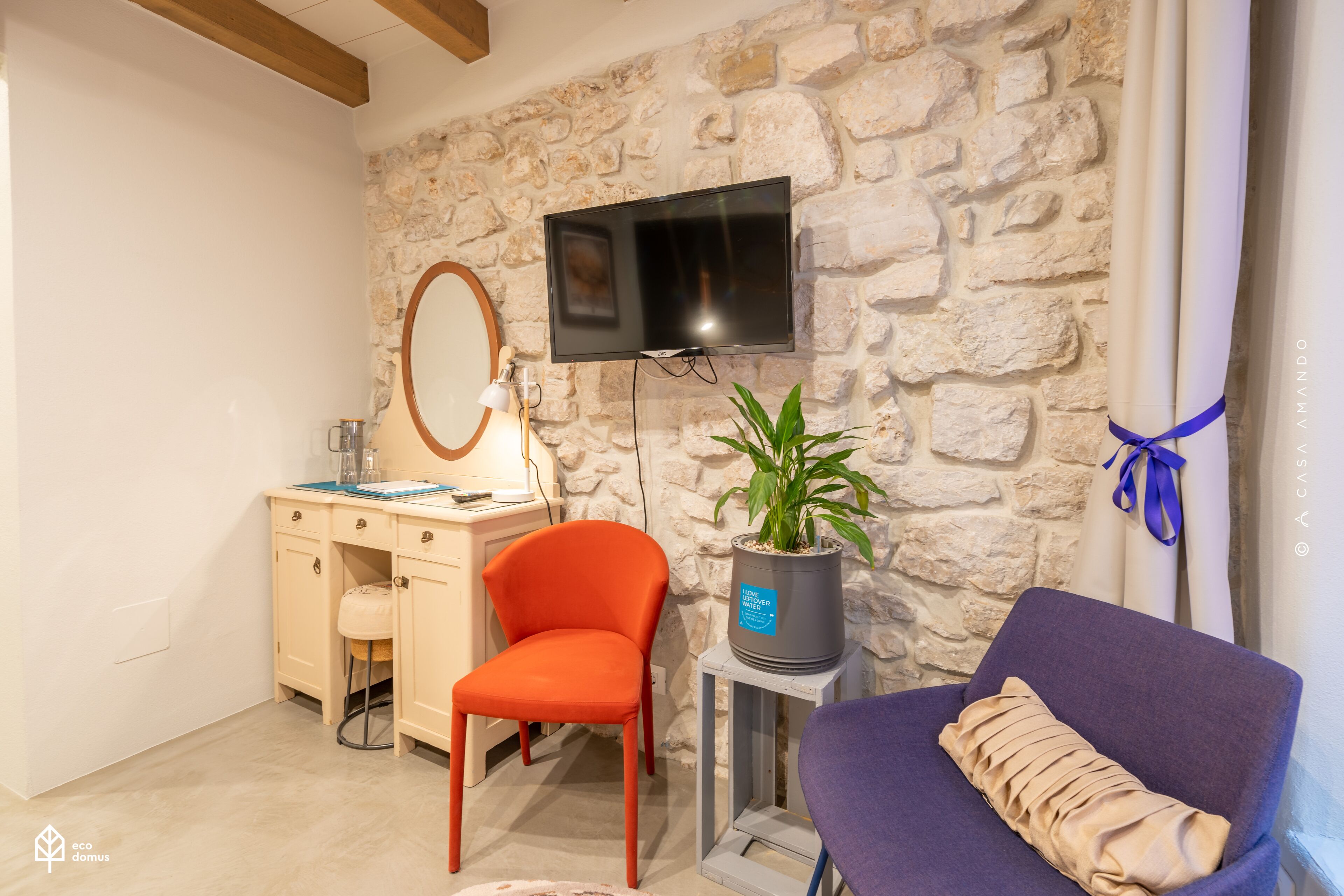 A room with a stone wall, a flat-screen TV, a desk with a chair, a small table with a potted plant, and a purple armchair.