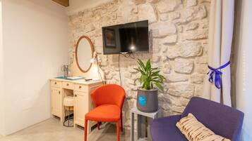 A room with a stone wall, a flat-screen TV, a desk with a chair, a small table with a potted plant, and a purple armchair.