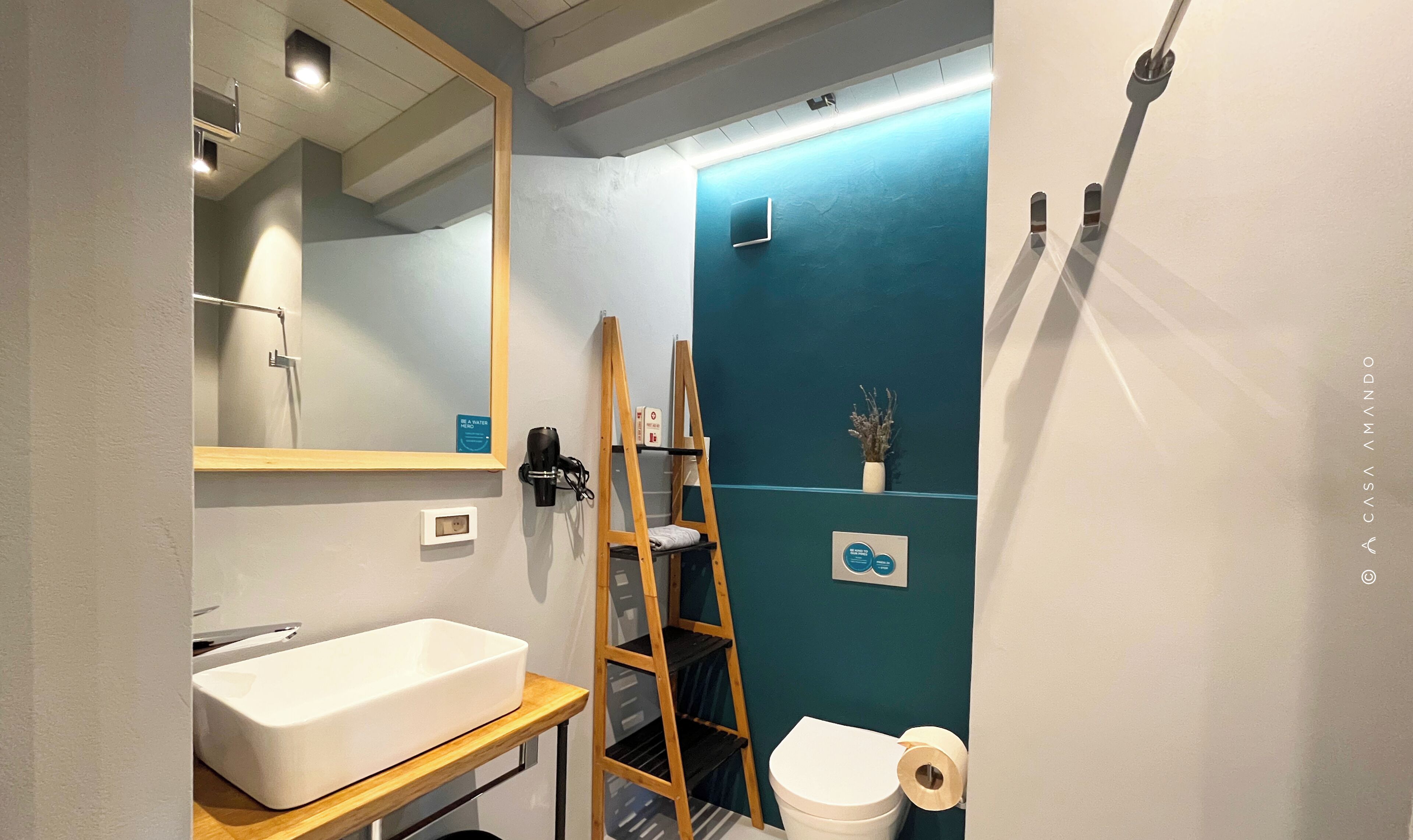 Un baño con un lavamanos blanco, un espejo, una escalera de madera, un inodoro y una pared azul.