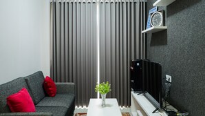 A living room with a grey sofa, red cushions, a white coffee table with a green plant, a black TV stand, and a clock on a shelf.