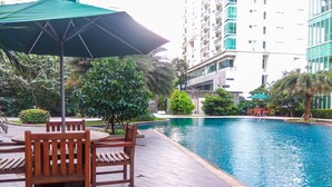 A poolside area with wooden furniture and a green umbrella, surrounded by lush greenery and modern buildings. - New Furnished Studio Apartment @ Woodland Park Residence (Jakarta)
