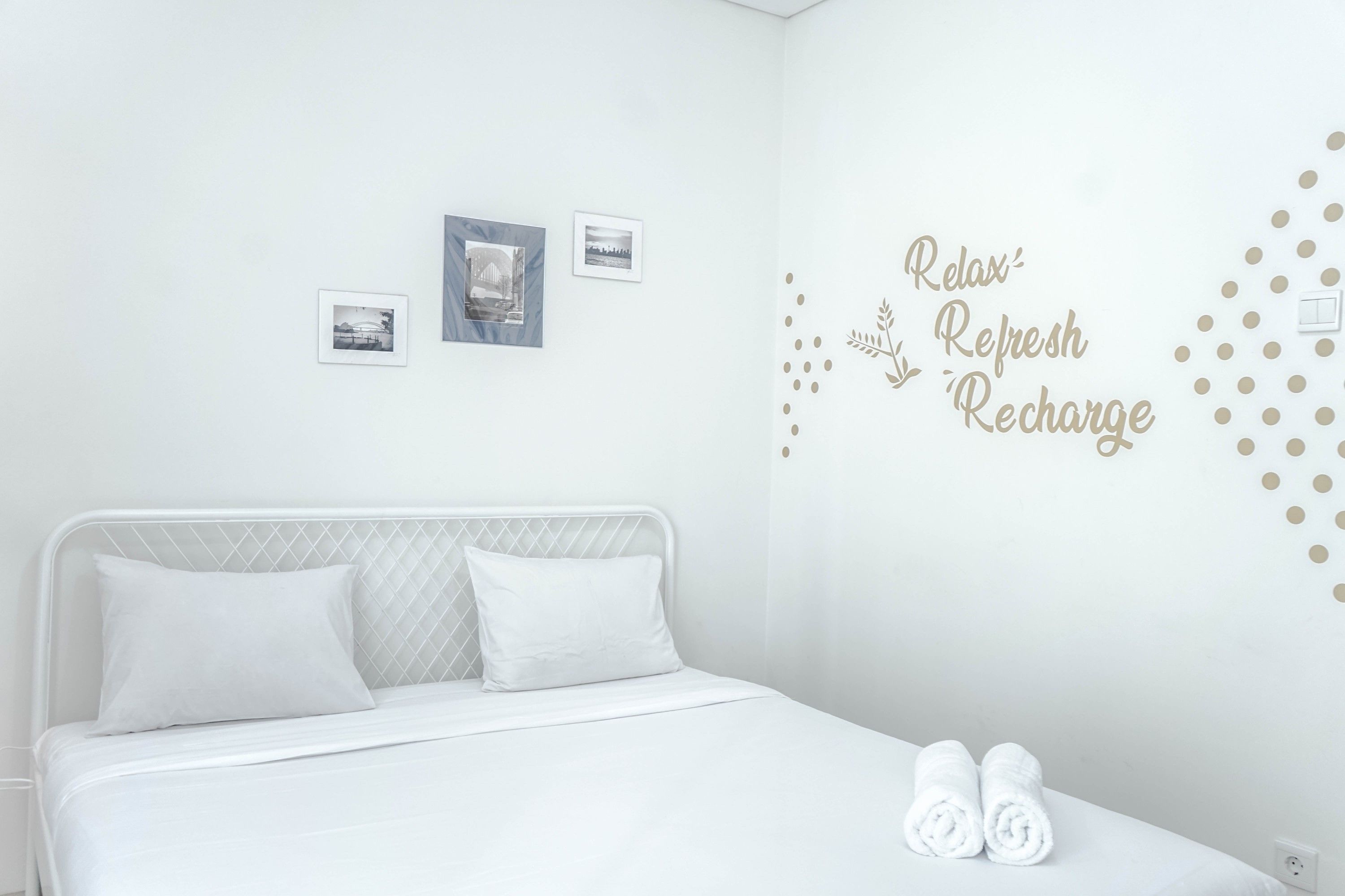 A clean, white hotel room with a bed, two pillows, and a wall with a 'Relax, Refresh, Recharge' sign.