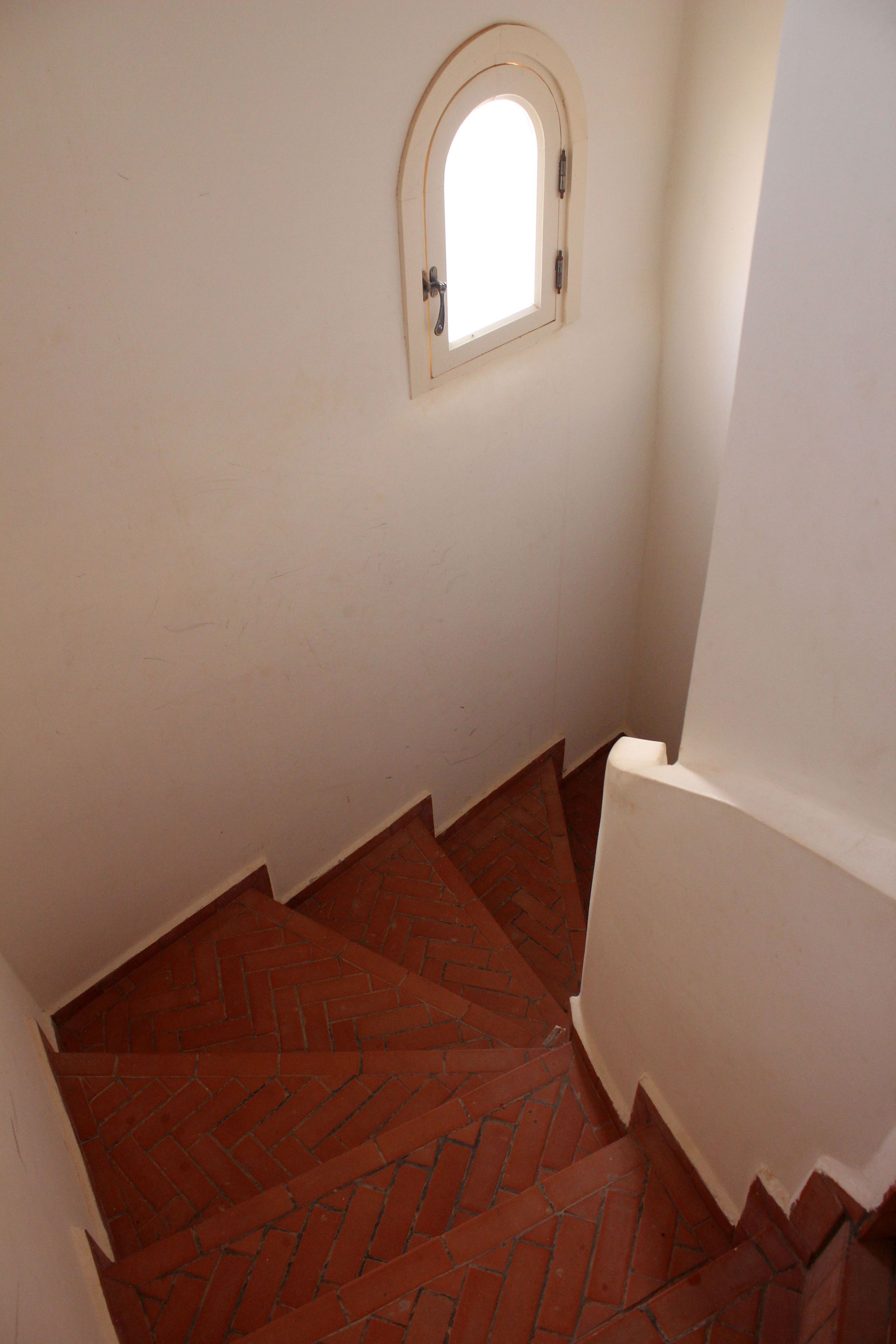Un escalier dont les marches sont recouvertes de briques rouges et les murs peints en blanc, menant à une porte surmontée d’une petite fenêtre.