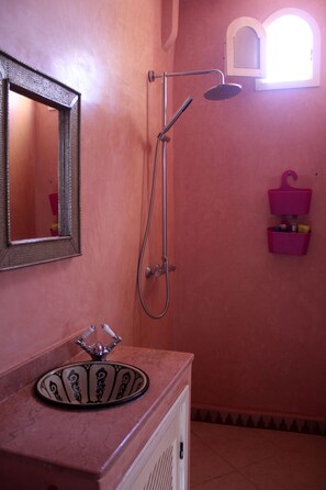 A bathroom with a pink wall, a round sink with a decorative basin, a mirror, a shower with a handheld showerhead, and a small window above the shower.