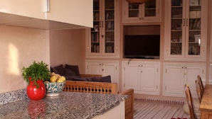 Een keuken met witte kastjes, een granieten aanrecht, een rode vaas met een plant, een geweven lamp en een eethoek met een houten tafel en stoelen.