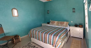 A bedroom with a teal wall, a bed with a striped blanket, a small wooden chair, a nightstand with a bowl, and a window with curtains.
