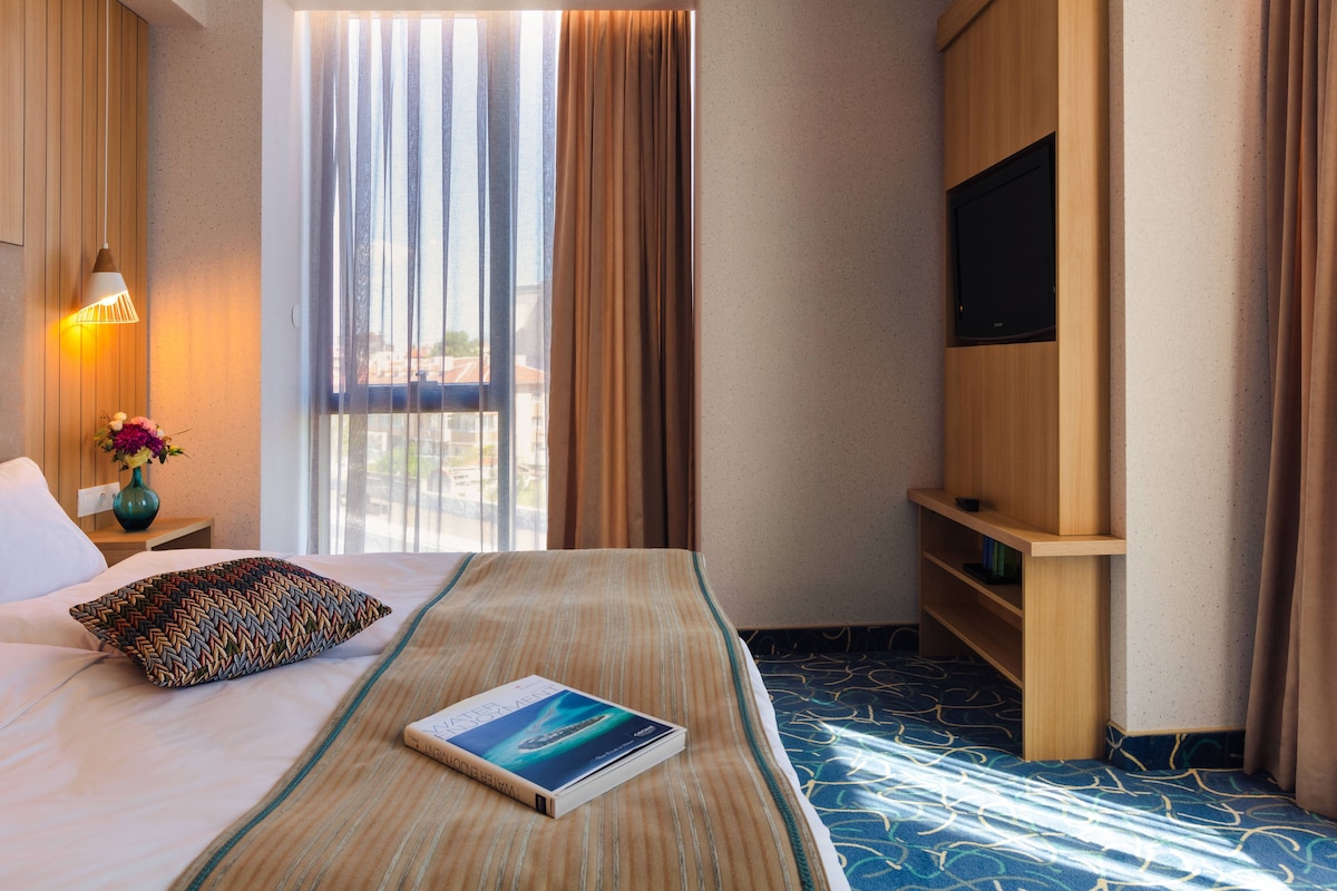 Une chambre d’hôtel avec un lit, une télévision, une bibliothèque, une fenêtre avec des rideaux et une lampe.