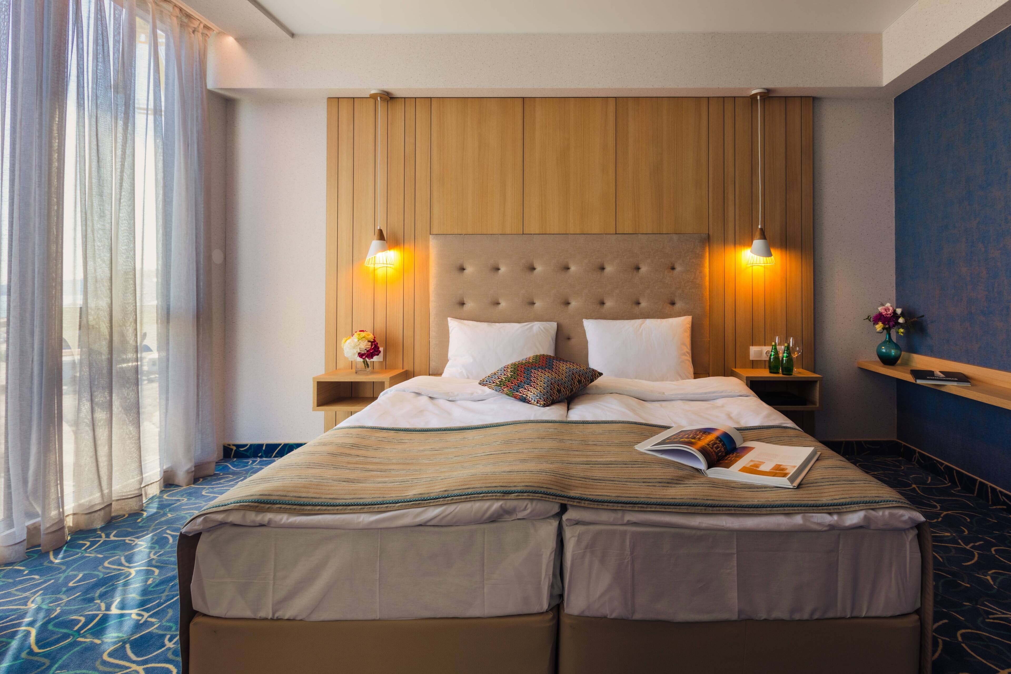A hotel room with a large bed, two bedside lamps, a nightstand with a book and a vase, and a view of the outdoors through sheer curtains.