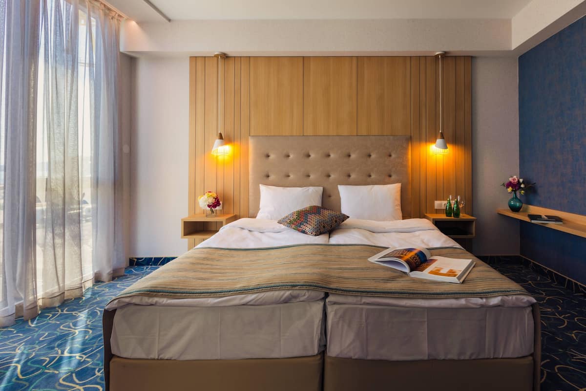 A hotel room with a large bed, two bedside lamps, a nightstand with a book and a vase, and a view of the outdoors through sheer curtains.