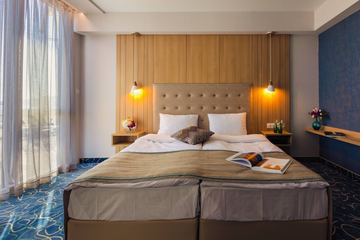 A hotel room with a large bed, two bedside lamps, a nightstand with a book and a vase, and a view of the outdoors through sheer curtains.