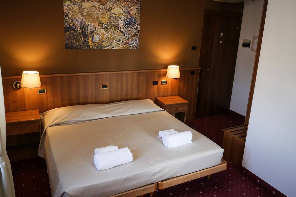 A hotel room with a large bed, two bedside lamps, a wooden headboard, a wall-mounted painting, and a red carpeted floor.