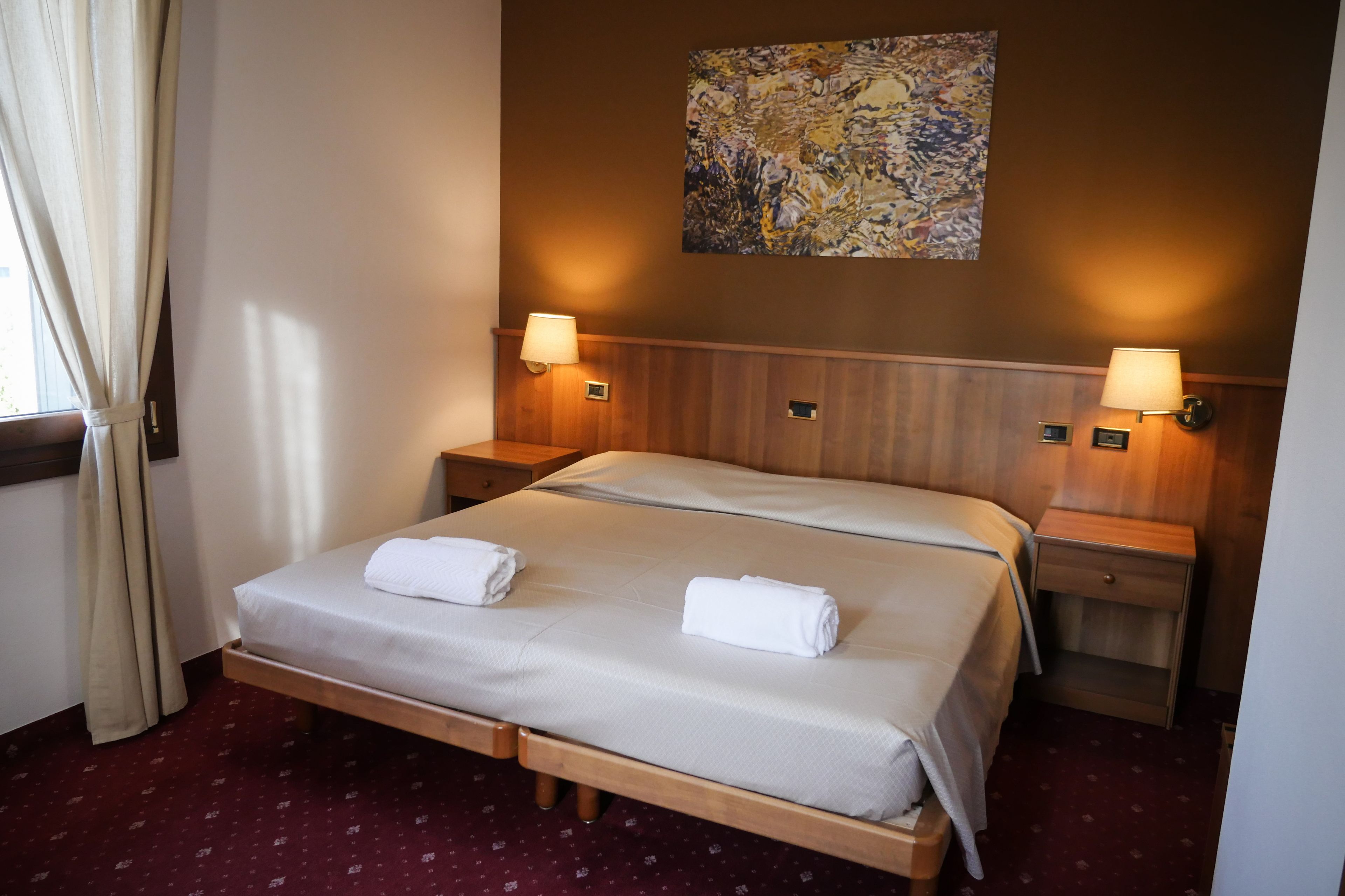 Habitación de hotel con una cama, dos mesitas de noche, un cuadro en la pared y una ventana con cortinas.