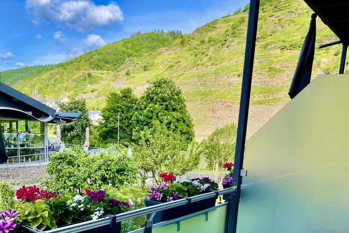 A balcony with a table and chairs, overlooking a green hillside with trees and flowers.