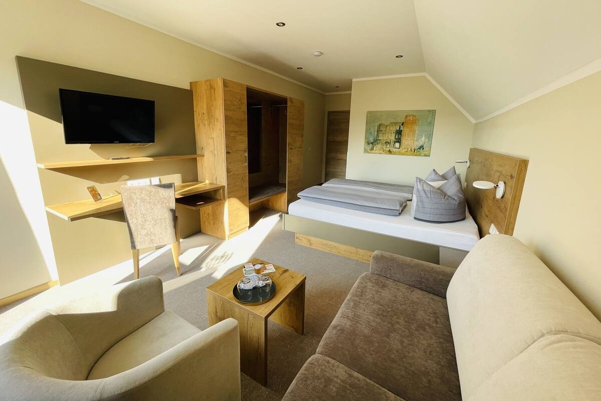 A modern, well-lit room with a flat-screen TV, a bed with gray bedding, a wooden desk, a chair, and a sofa.