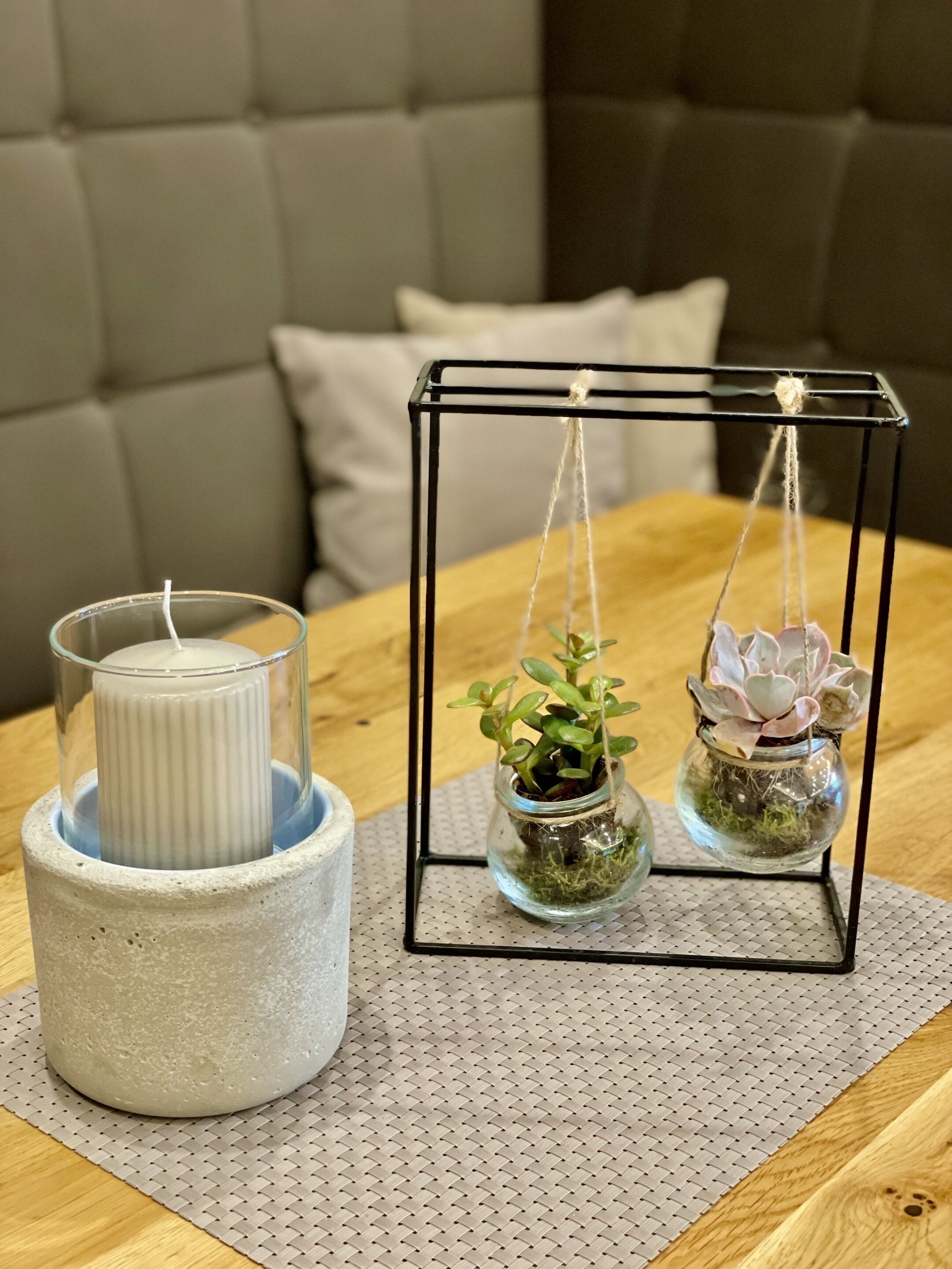 Una vela en un portaluz decorativo sobre una mesa de madera, con dos frascos de vidrio colgantes con plantas.