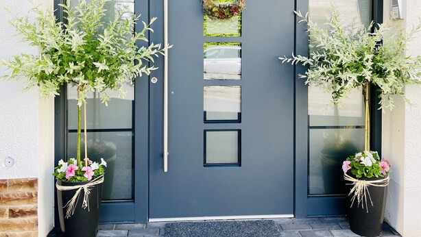 Una puerta gris oscuro con un picaporte vertical y un pequeño adorno floral encima. A cada lado de la puerta hay dos macetas con follaje verde y flores rosadas.