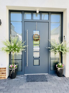 A dark grey door with a vertical handle and a small wreath above it. There are two potted plants with green foliage and pink flowers on either side of the door. - Weinbergs Loge (Ernst)