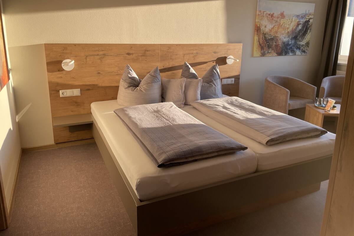 A hotel room with a wooden headboard, two beds, a chair, a small table, and a painting on the wall.