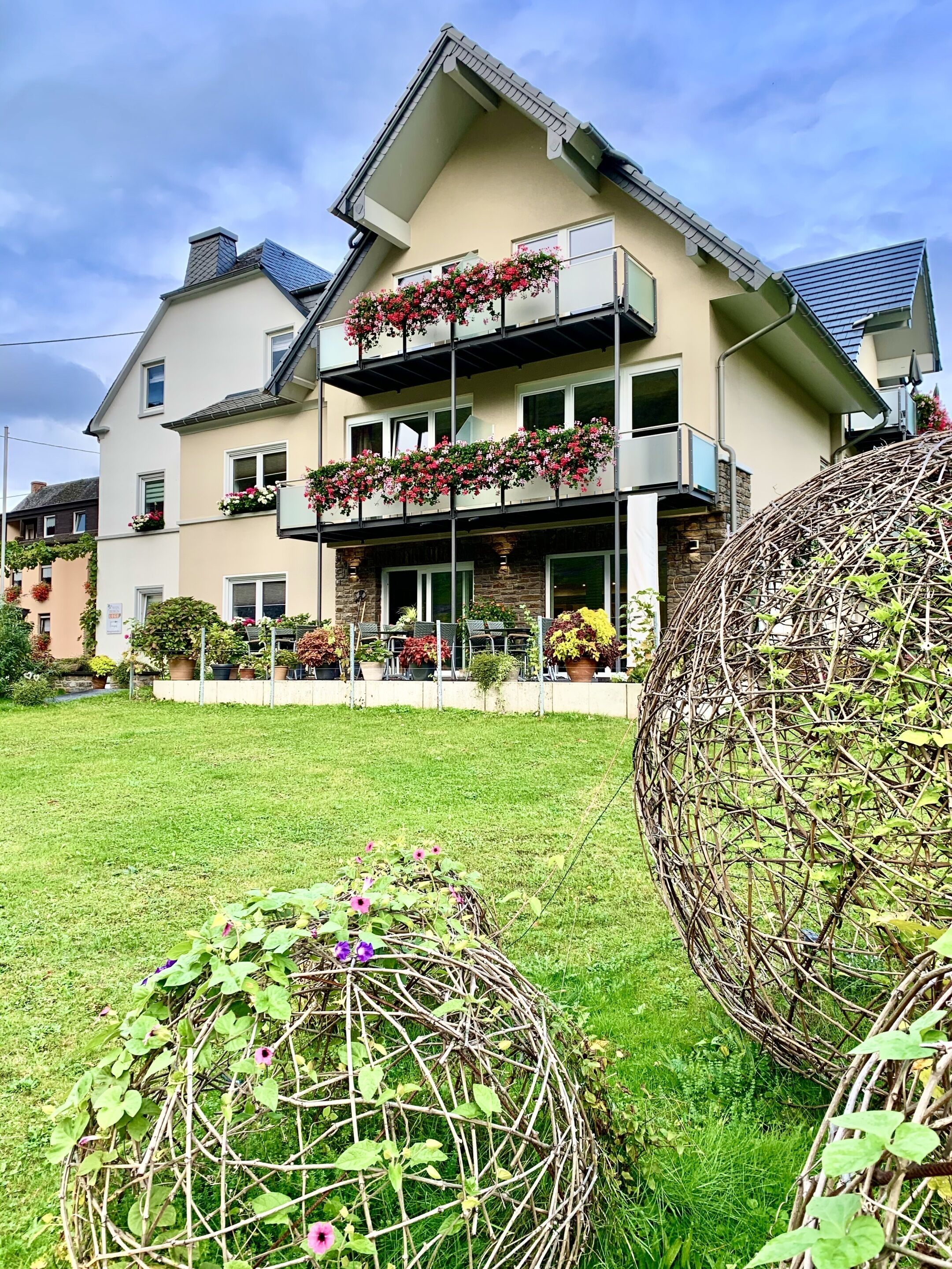 Ein zweistöckiges Haus mit einem Balkon, der mit Blumen geschmückt ist, einem gepflegten Rasen und dekorativen, kugelförmigen Elementen.