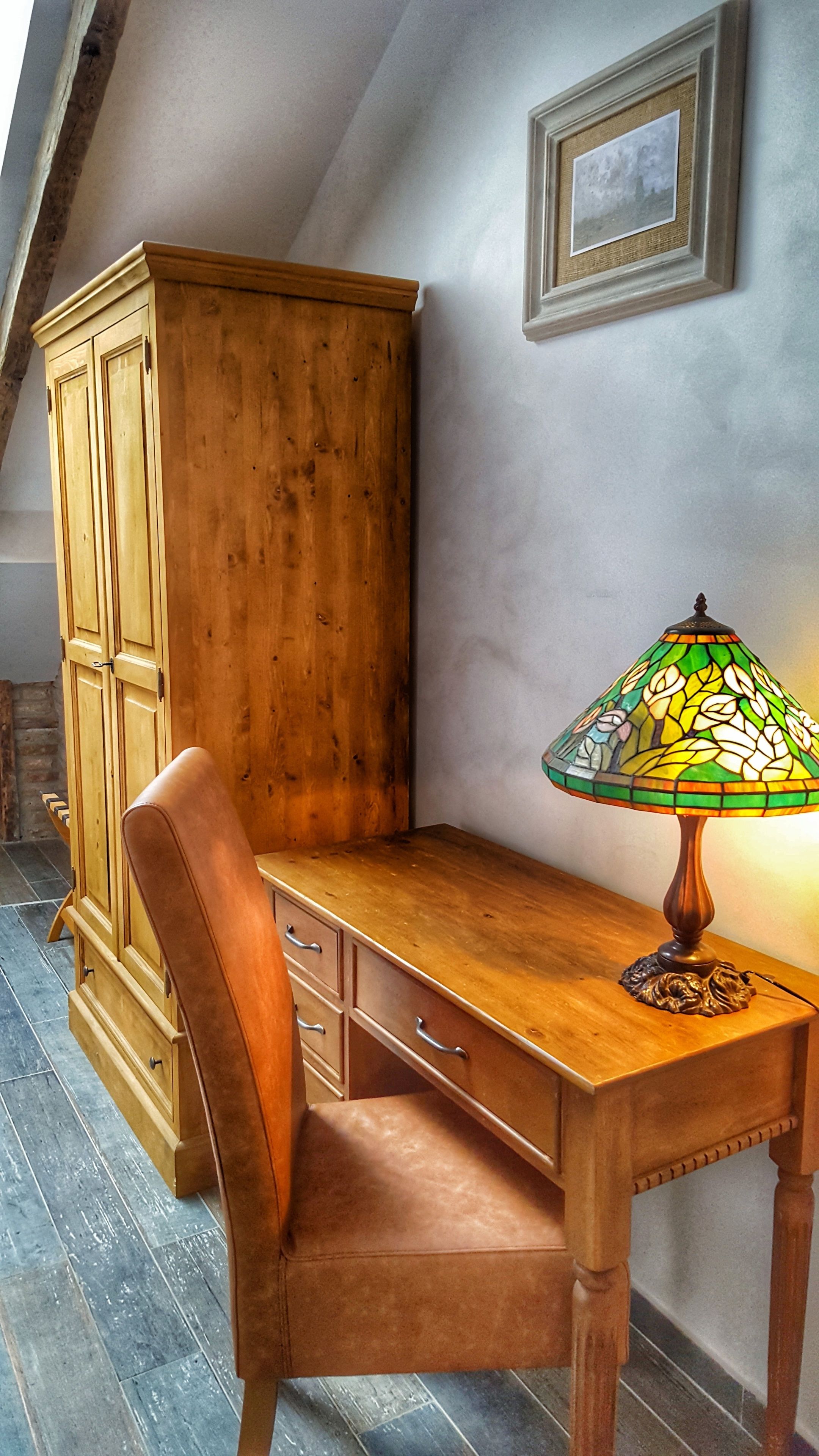 A wooden desk with a stained glass lamp, a leather chair, and a wooden cabinet in a room with sloped ceilings and a tiled floor.