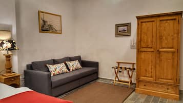 Um quarto com um sofá cinzento, duas almofadas coloridas, um guarda-roupa de madeira, uma mesa pequena de madeira e uma luminária num criado-mudo.