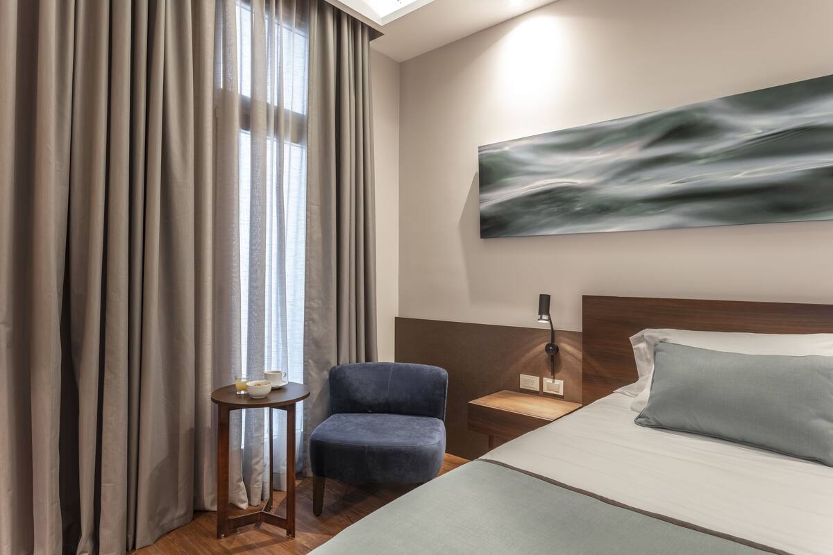 A hotel room with a bed, a chair, a small table with a lamp, and a painting on the wall.