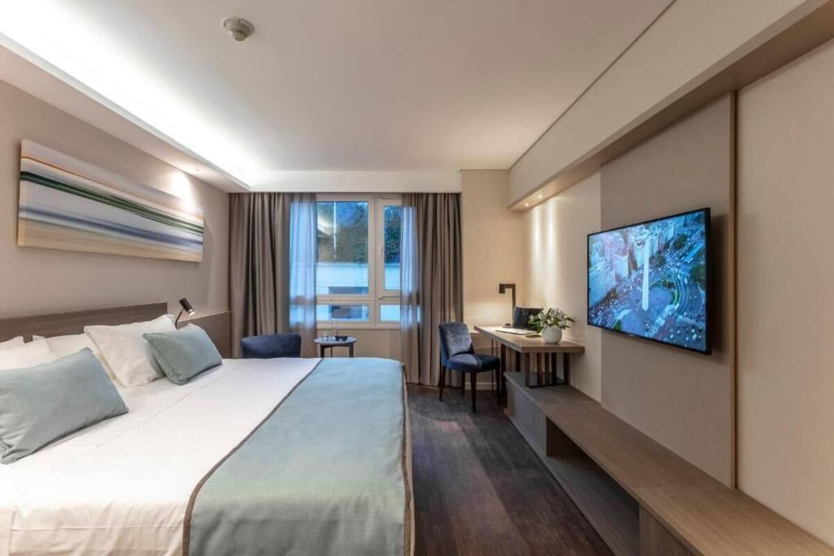 A hotel room with a bed, a desk, a chair, a TV, and a window with curtains.