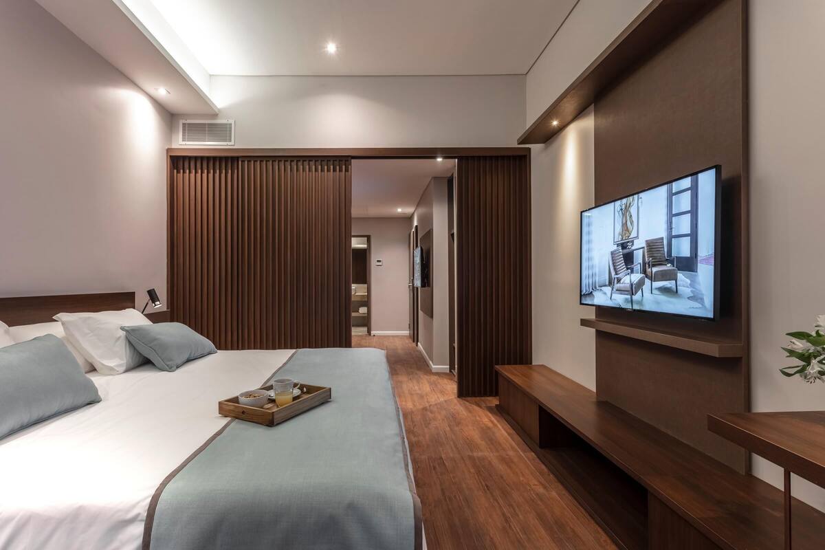 A modern hotel room with a large bed, a flat-screen TV, wooden paneling, and a wooden bench.