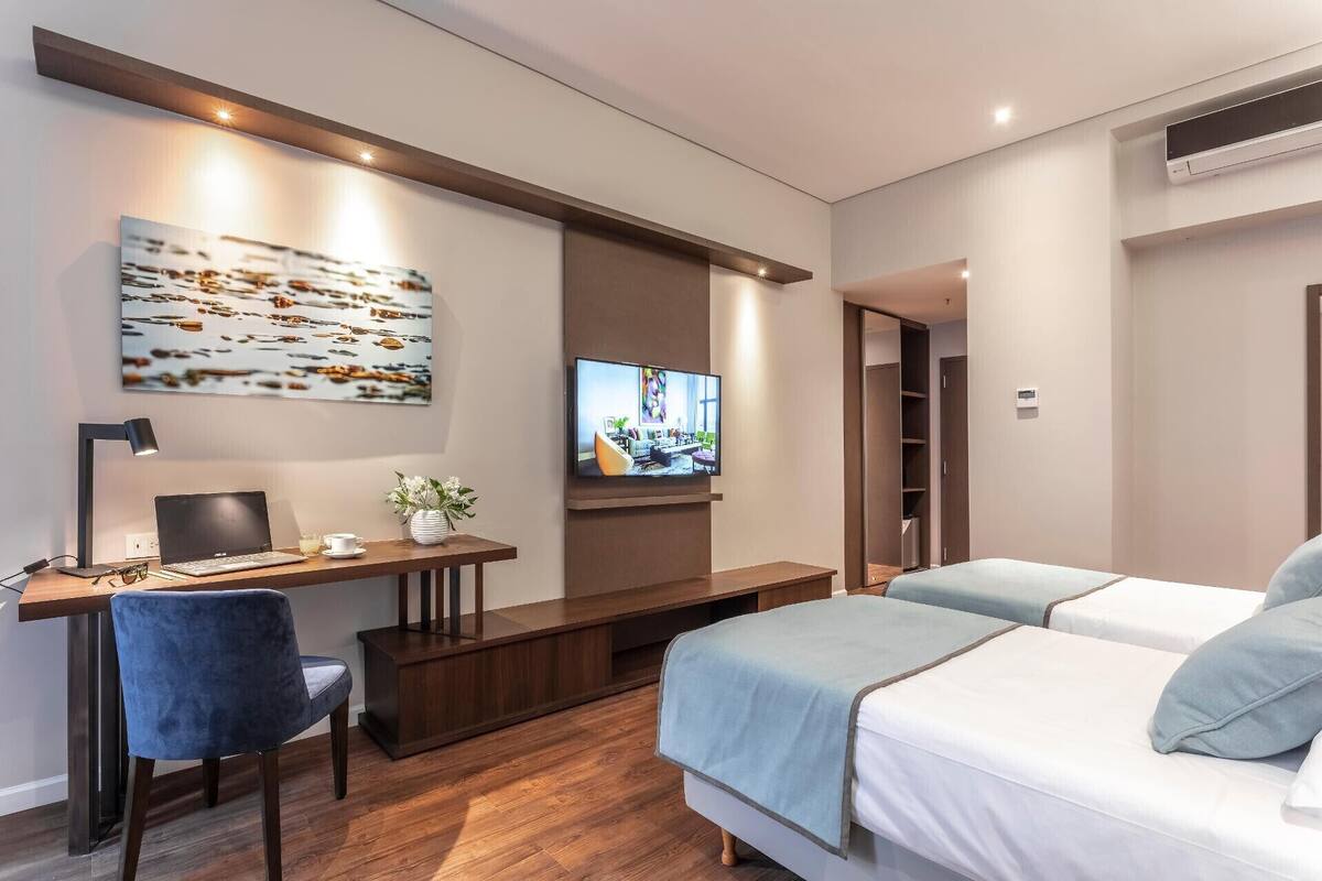 A modern hotel room with a desk, laptop, chair, TV, and a bed with two pillows.