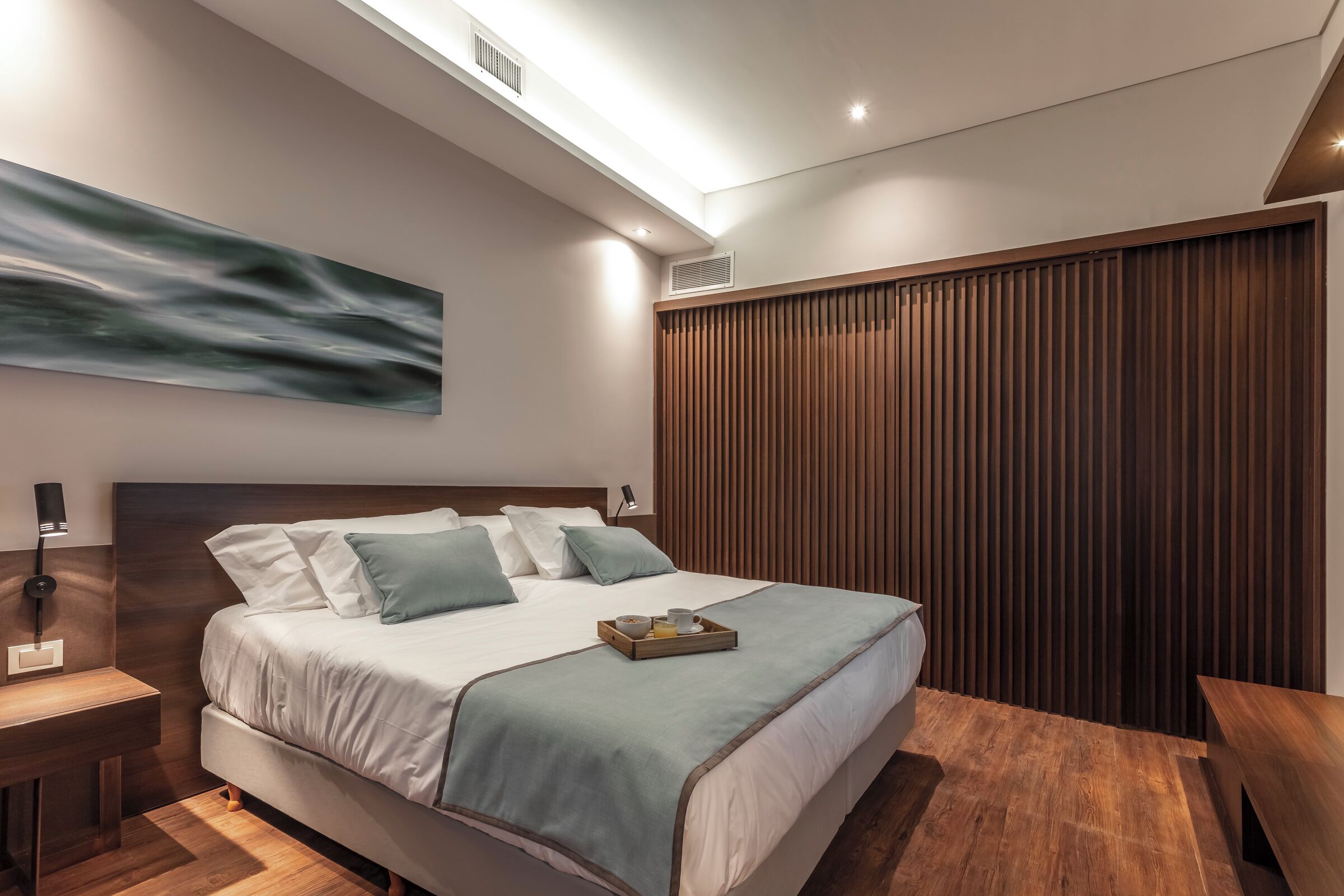 A modern bedroom with a bed, bedside tables, a wall-mounted artwork, and a wooden slatted wall.