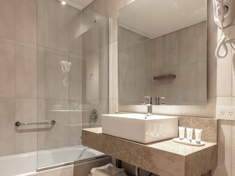 Une salle de bain moderne dotée d’une douche à parois de verre, d’un lavabo blanc avec un plan de toilette en marbre, d’un grand miroir et d’un sèche-cheveux fixé au mur.