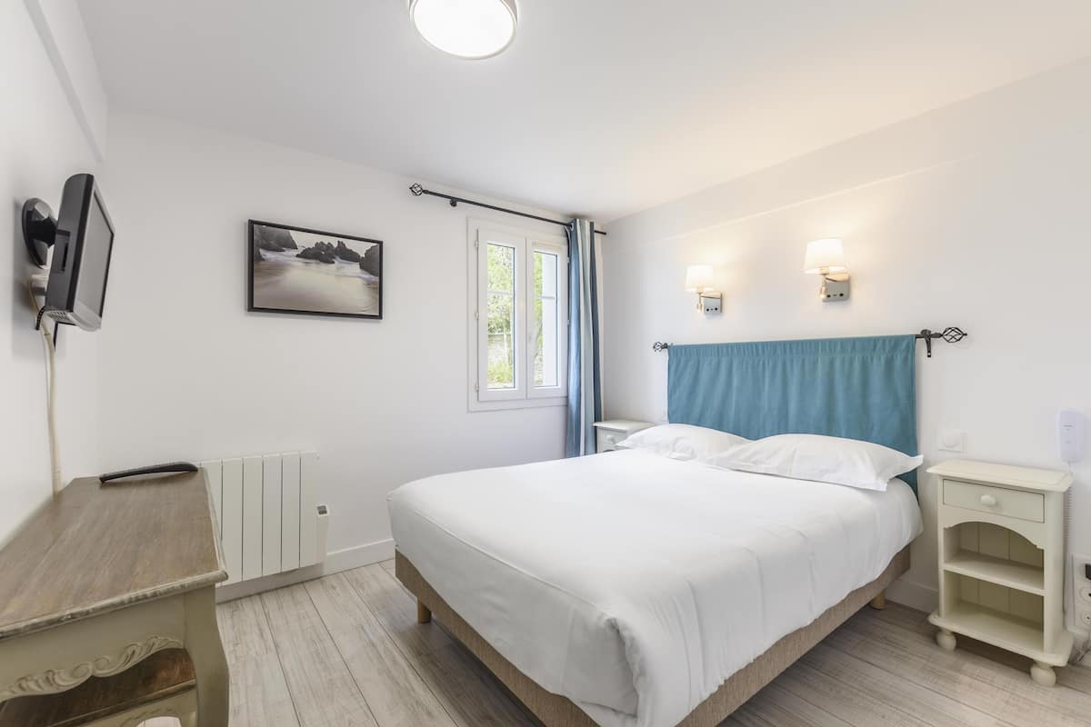 A bedroom with a bed, a bedside table, a nightstand, a wall-mounted TV, a window with curtains, and a picture on the wall.