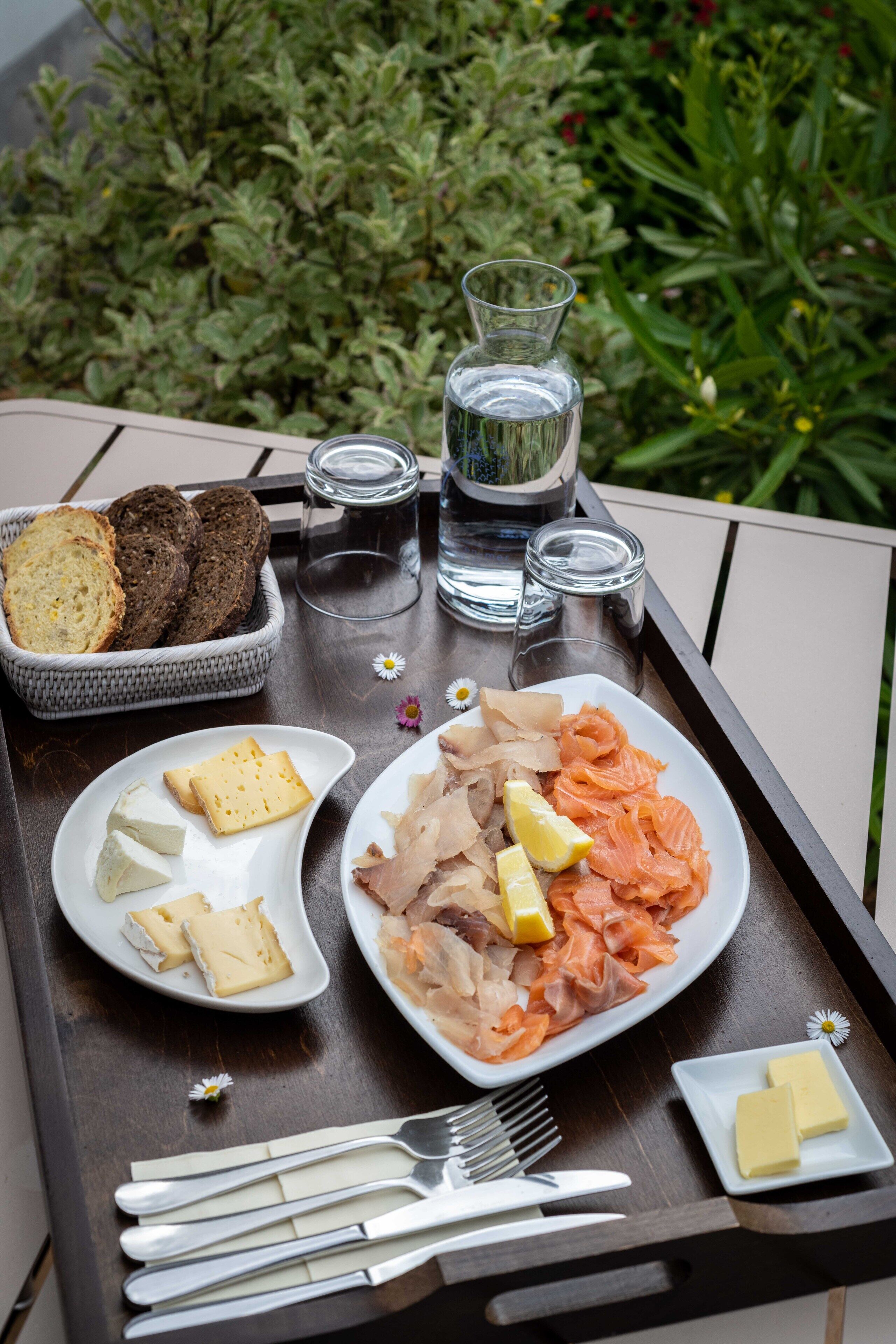 A tray with food items including salmon, cheese, and bread, accompanied by butter and utensils, set on an outdoor table with a garden view.
