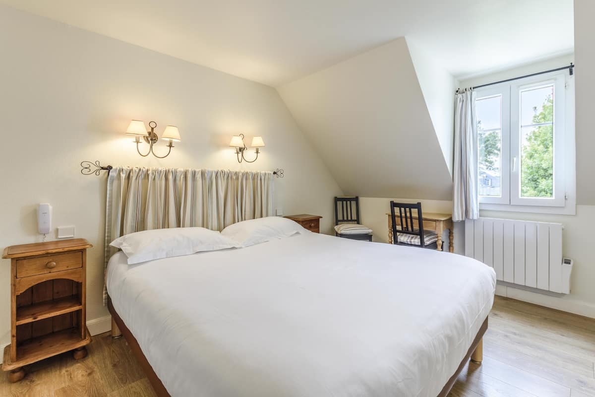 A bedroom with a large bed, two bedside tables, a nightstand, a chair, a window with curtains, and wall-mounted lamps.