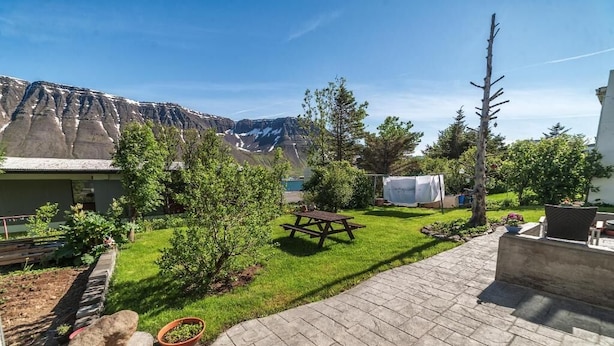 A backyard with a picnic table, a stone bench, a small garden, and a view of a mountain range. - Sólheimar Studio Apartments (Isafjordur)