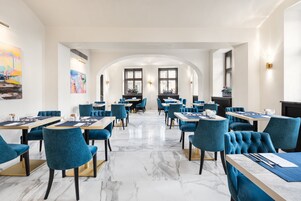 Uma sala de jantar moderna com piso de mármore, cadeiras estofadas em azul e mesas dispostas em um layout espaçoso.