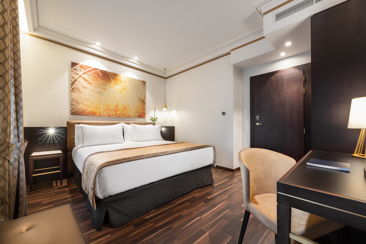 A hotel room with a large bed, a desk, a chair, a painting on the wall, and a door.