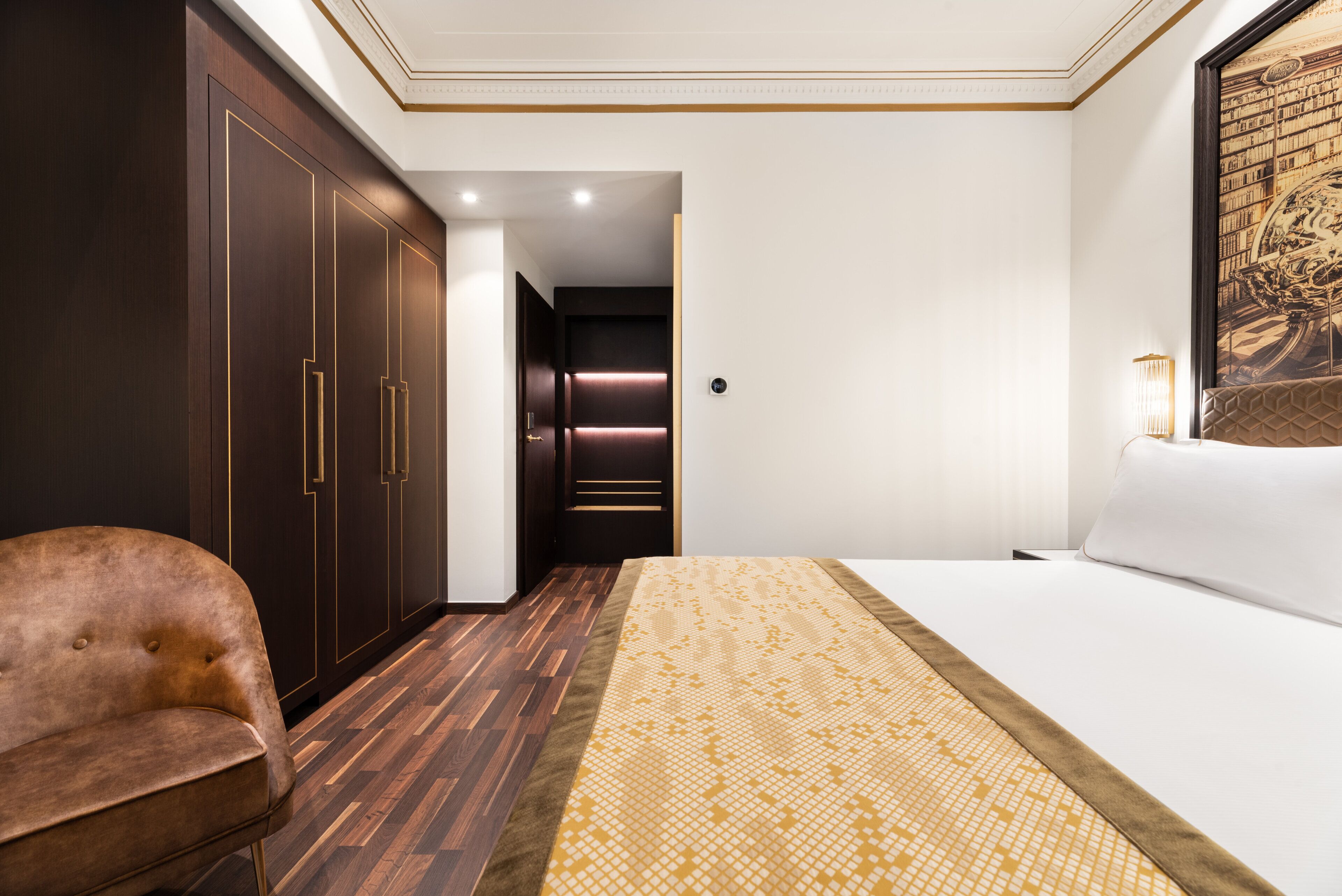 A hotel room with a large bed, a brown leather chair, dark wooden wardrobes, and a wall-mounted lamp.