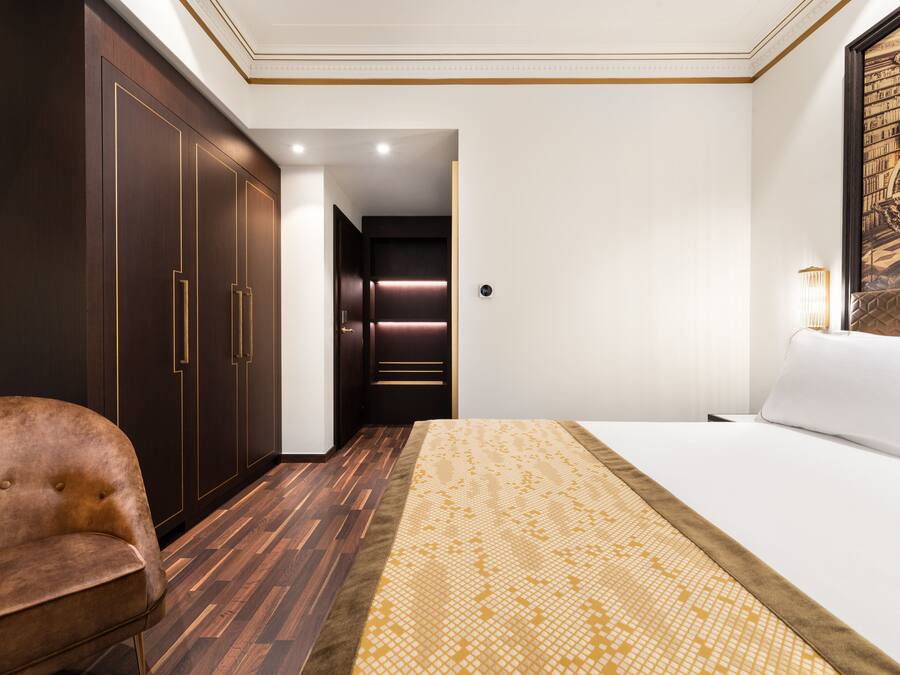 A hotel room with a large bed, a brown leather chair, dark wooden wardrobes, and a wall-mounted lamp.