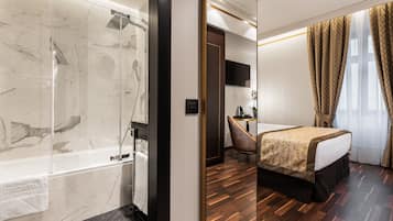 A hotel room with a marble bathroom, a wooden floor, a bed with a gold and brown bedspread, a desk, and a TV mounted on the wall.