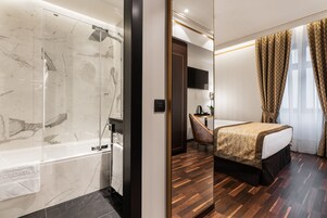 Quarto de hotel com casa de banho em mármore, chão de madeira, cama com colcha dourada e castanha, secretária e televisão na parede.