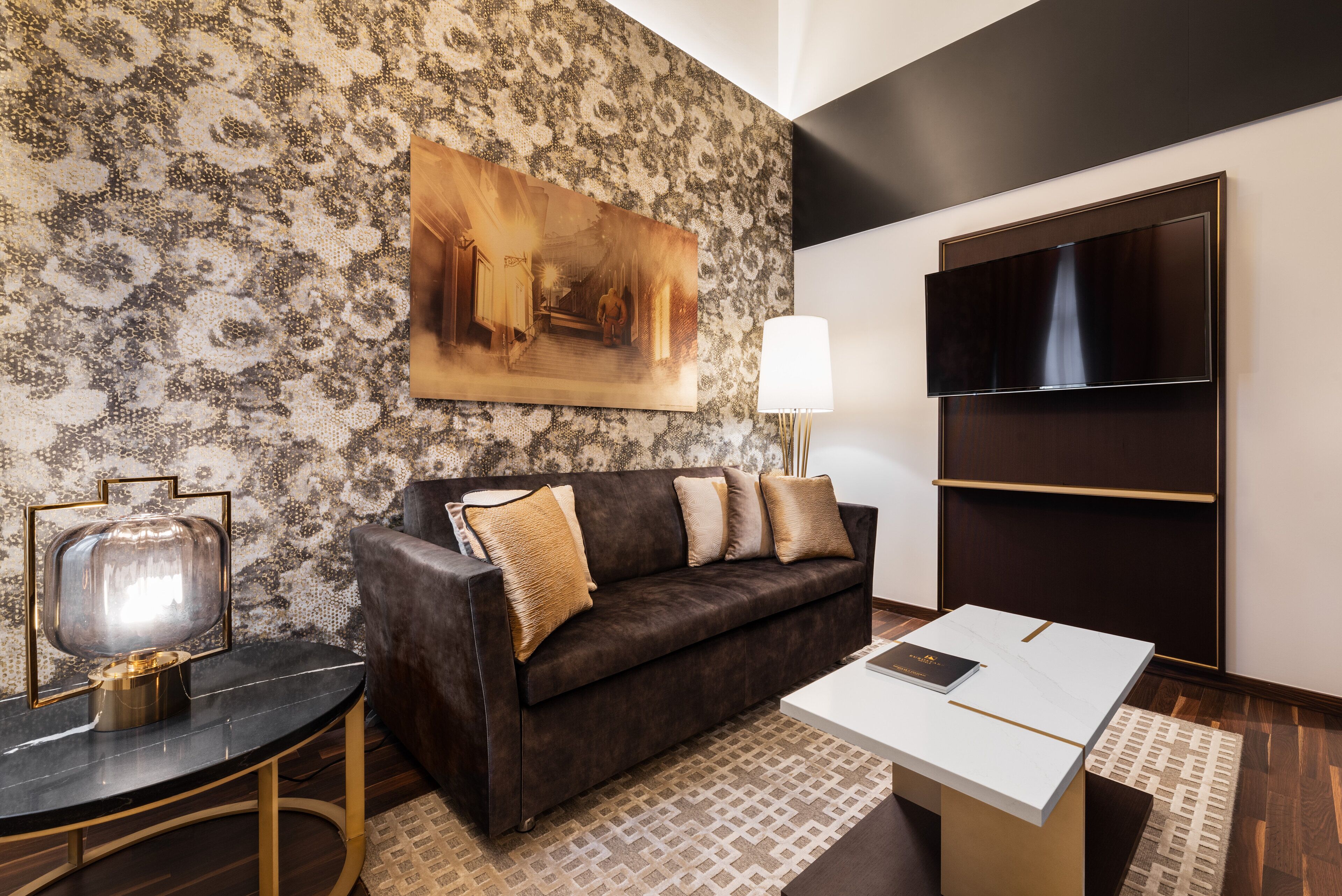 A modern living room with a patterned wall, a dark brown sofa with cushions, a flat-screen TV, a table with a lamp, and a framed painting.