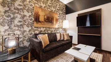 A modern living room with a patterned wall, a dark brown sofa with cushions, a flat-screen TV, a table with a lamp, and a framed painting.